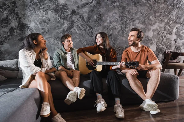 Donna allegra che suona la chitarra acustica vicino agli amici multietnici seduti sul divano in soggiorno moderno — Foto stock