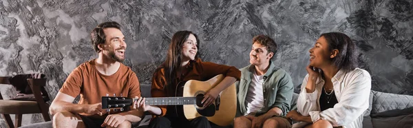 Mulher alegre tocando guitarra acústica perto de amigos multiétnicos sentados no sofá na sala de estar moderna, banner — Fotografia de Stock