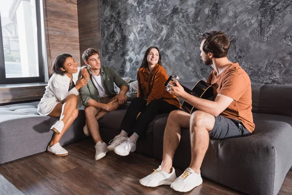 Homem tocando guitarra acústica perto de amigos multiétnicos felizes sentados no sofá na sala de estar moderna — Fotografia de Stock