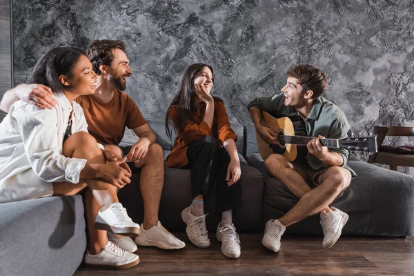 Aufgeregter Mann spielt Akustikgitarre in der Nähe fröhlicher multiethnischer Freunde, die auf der Couch im modernen Wohnzimmer sitzen — Stockfoto