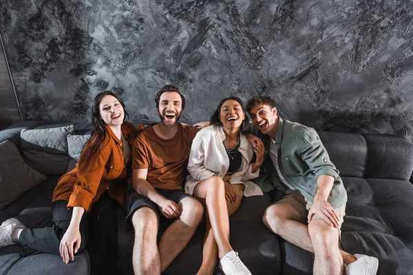 Cheerful multicultural friends in casual clothing sitting on grey couch in modern living room — Stock Photo