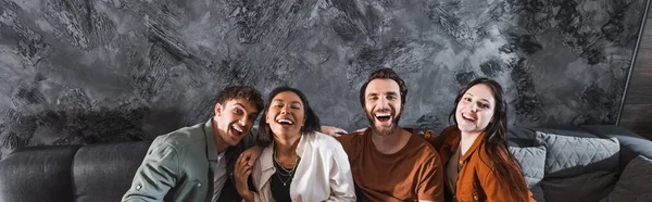 Fröhliche multikulturelle Freunde in lässiger Kleidung auf grauer Couch im modernen Wohnzimmer sitzend, Banner — Stockfoto