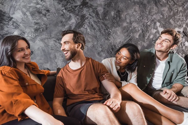 Amigos multiculturais positivos em roupas casuais sentados no sofá cinza na sala de estar moderna — Fotografia de Stock