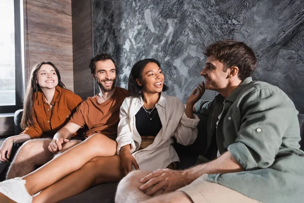 Amigos multiculturales felices en ropa casual sentado en el sofá gris en la sala de estar moderna - foto de stock