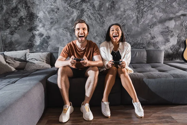 KYIV, UCRANIA - 26 de julio de 2022: amigos interracial sorprendidos jugando videojuegos y sentados en un sofá gris - foto de stock