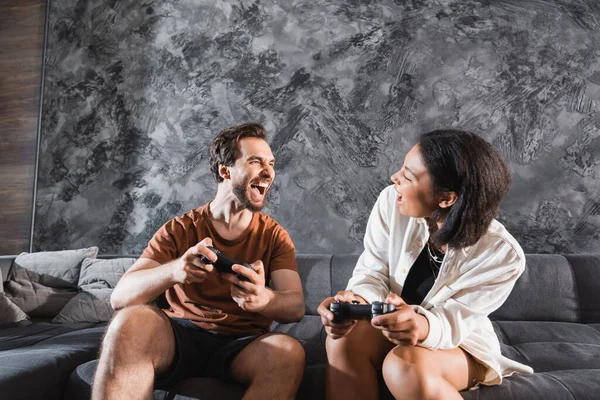 KYIV, UKRAINE - JULY 26, 2022: happy interracial friends holding joysticks and looking at each other — Stock Photo