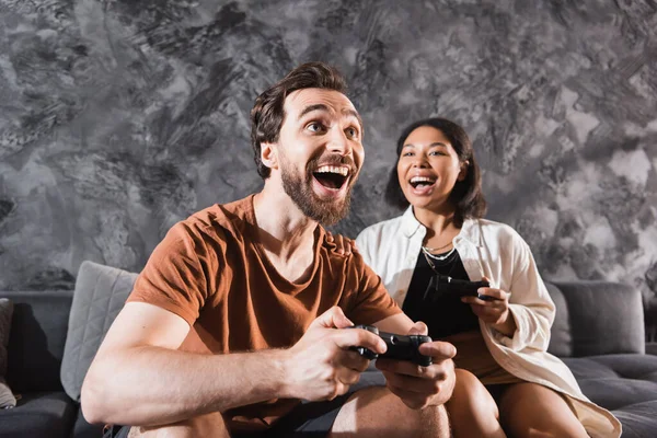 KYIV, UCRANIA - 26 de julio de 2022: hombre excitado sosteniendo el joystick y jugando videojuegos con mujeres bi-raciales en la sala de estar - foto de stock