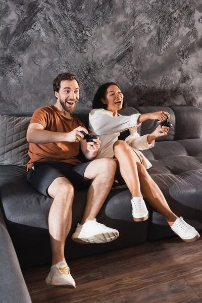 KYIV, UCRANIA - 26 de julio de 2022: felices amigos multiétnicos jugando videojuegos y sentados en un sofá gris - foto de stock