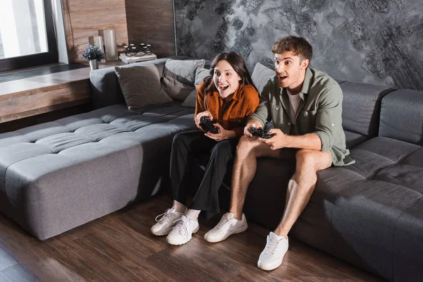KYIV, UCRANIA - 26 de julio de 2022: amigos emocionados sosteniendo joysticks y jugando videojuegos mientras están sentados en un sofá gris - foto de stock