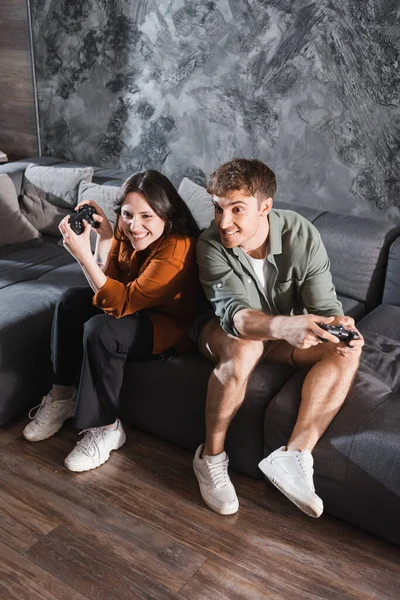 KYIV, UCRANIA - 26 de julio de 2022: amigos alegres sosteniendo joysticks y jugando videojuegos mientras están sentados en un sofá gris - foto de stock