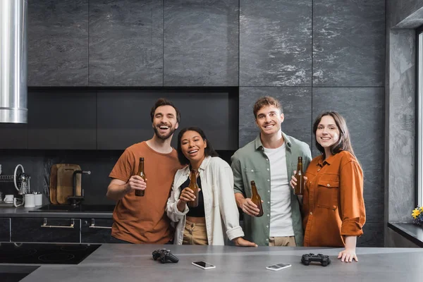 KYIV, UKRAINE - JULY 26, 2022: happy interracial friends holding bottles of beer near joysticks and smartphones on kitchen worktop — Stock Photo