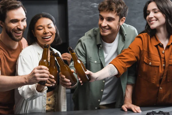 KYIV, UKRAINE - JULY 26, 2022: happy interracial friends clinking bottles of beer near joystick on kitchen worktop — Stock Photo