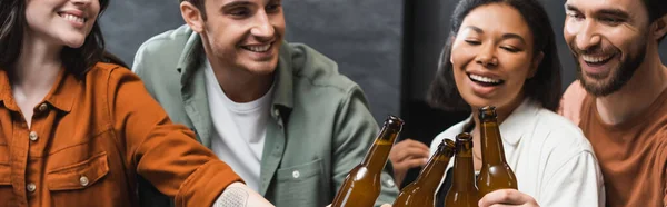 Heureux interracial amis souriant et cliquetis bouteilles de bière dans la cuisine, bannière — Photo de stock