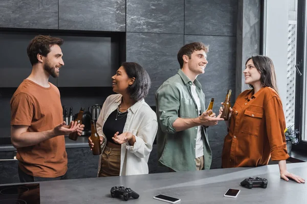 QUIIV, UCRÂNIA - JULHO 26, 2022: amigos inter-raciais felizes conversando enquanto seguram garrafas de cerveja perto de gadgets e joysticks na bancada da cozinha — Fotografia de Stock