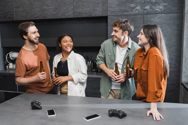 KYIV, UKRAINE - JULY 26, 2022: smiling interracial friends holding bottles of beer near joysticks and smartphones on kitchen worktop — Stock Photo