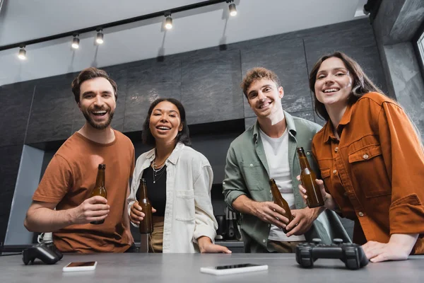 KYIV, UCRANIA - 26 de julio de 2022: amigos multiétnicos sonrientes sosteniendo botellas de cerveza cerca de joysticks y teléfonos inteligentes en la encimera de la cocina - foto de stock