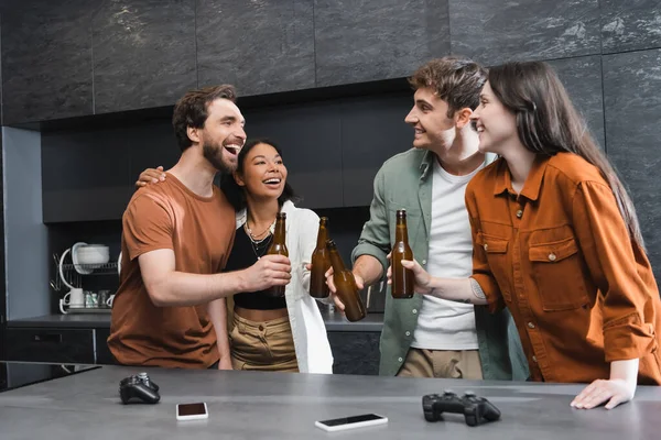 KYIV, UKRAINE - JULY 26, 2022: cheerful interracial friends clinking bottles of beer near joysticks on kitchen worktop — Stock Photo