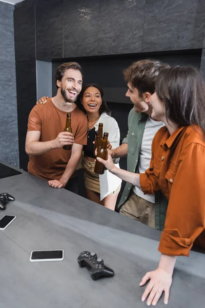 KYIV, UKRAINE - JULY 26, 2022: cheerful multiethnic friends clinking bottles of beer near joysticks on kitchen worktop — Stock Photo