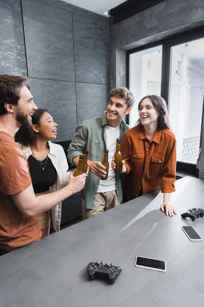 KYIV, UKRAINE - JULY 26, 2022: cheerful multiethnic friends holding bottles of beer near joysticks on kitchen worktop — Stock Photo