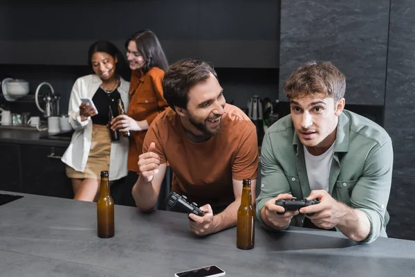 KYIV, UKRAINE - JULY 26, 2022: cheerful men playing video game near blurred interracial women using smartphone in kitchen — Stock Photo