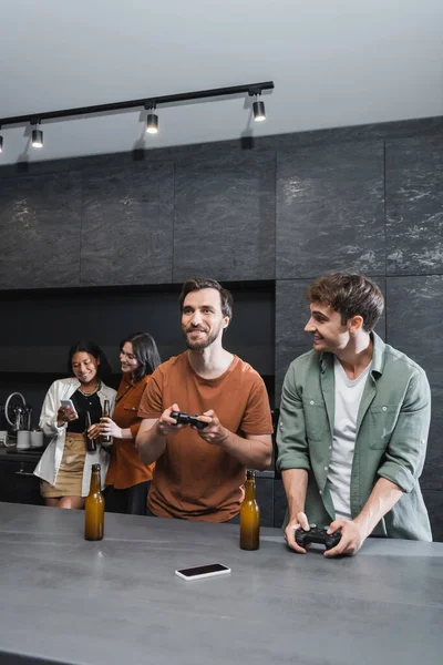 KYIV, UCRANIA - 26 de julio de 2022: hombres felices jugando videojuegos cerca de mujeres interracial usando teléfonos inteligentes en la cocina - foto de stock