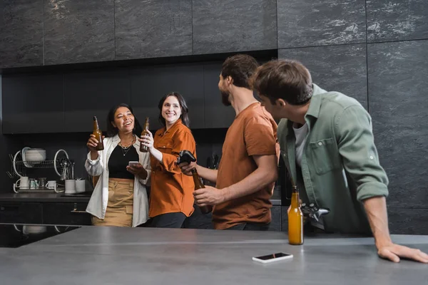 KYIV, UCRANIA - 26 de julio de 2022: hombres con joysticks mirando a mujeres interracial felices sosteniendo botellas de cerveza en la cocina - foto de stock