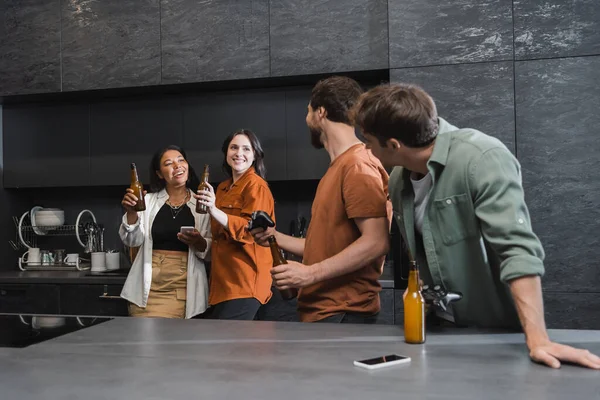 KYIV, UKRAINE - 26 JUILLET 2022 : hommes avec des joysticks regardant des femmes interraciales heureuses tenant des bouteilles de bière dans la cuisine — Photo de stock