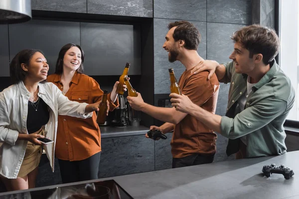 KYIV, UKRAINE - JULY 26, 2022: cheerful multiethnic friends clinking bottles of beer near joysticks in kitchen — Stock Photo