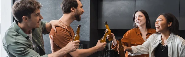 Amigos multiétnicos felizes sorrindo e batendo garrafas de cerveja na cozinha, banner — Fotografia de Stock