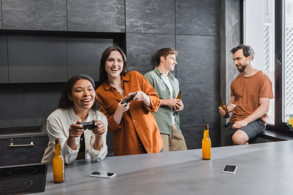 KYIV, UCRANIA - 26 de julio de 2022: felices mujeres interracial jugando videojuegos cerca de amigos en la cocina - foto de stock