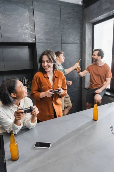 KYIV, UCRANIA - 26 de julio de 2022: alegres mujeres interracial jugando videojuegos cerca de amigos en la cocina — Stock Photo