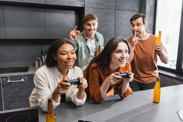 KYIV, UKRAINE - 26 JUILLET 2022 : joyeuses femmes interraciales jouant à un jeu vidéo près de l'homme avec de la bière montrant signe de silence dans la cuisine — Stock Photo