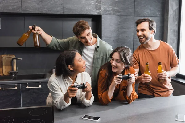 KYIV, UKRAINE - 26 JUILLET 2022 : joyeuses femmes interraciales jouant à un jeu vidéo près d'amis joyeux dans la cuisine — Stock Photo