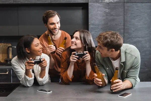 KYIV, UKRAINE - 26 JUILLET 2022 : joyeuses femmes interracial tenant des joysticks près d'amis masculins avec de la bière dans la cuisine — Photo de stock