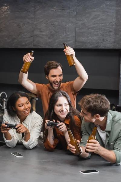 KIEW, UKRAINE - 26. JULI 2022: glückliche interrassische Frauen mit Steuerknüppeln in der Nähe von Freunden mit Bier in der Küche — Stockfoto