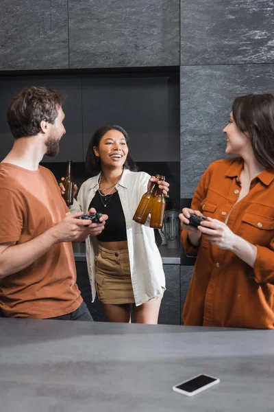 KYIV, UKRAINE - JULY 26, 2022: cheerful bi-racial woman giving beer to friends with joysticks in kitchen — Stock Photo