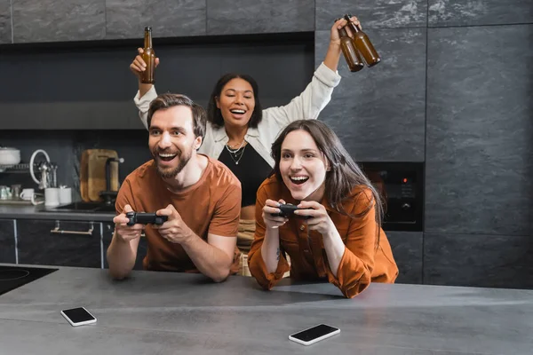 KYIV, UKRAINE - JULY 26, 2022: happy man and woman playing video game near excited bi-racial friend with bottles of beer — Stock Photo