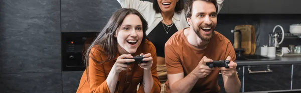 KYIV, UKRAINE - JULY 26, 2022: happy man and woman playing video game near excited bi-racial friend, banner — Stock Photo