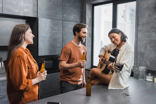 Щаслива бі-расова жінка грає на акустичній гітарі біля друзів з пляшками пива — стокове фото