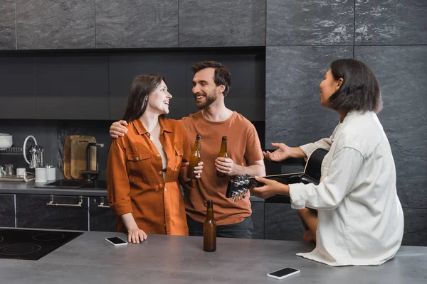 Homme heureux embrasser femme près bi-racial ami avec guitare acoustique geste dans la cuisine — Photo de stock