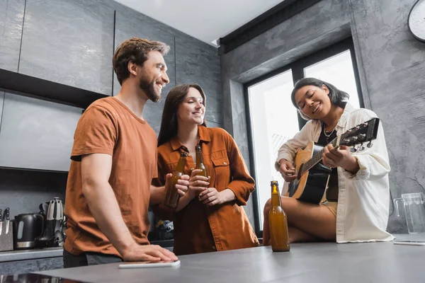 Low angle view of happy bi-racial woman playing acoustic guitar near cheerful friends with bottles of beer — Stock Photo