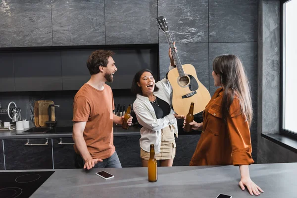 Femme bi-raciale étonnée tenant la guitare acoustique près des amis joyeux avec des bouteilles de bière — Photo de stock