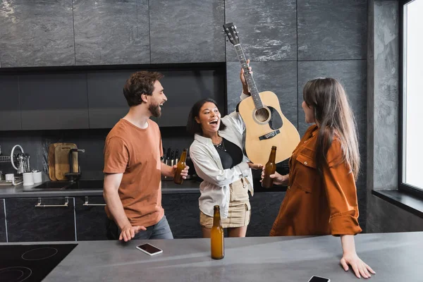 Amazed bi-racial woman holding  acoustic guitar near cheerful friends with bottles of beer — Stock Photo