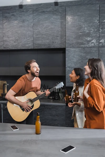 Homem alegre tocando guitarra acústica perto de mulheres inter-raciais felizes com garrafas de cerveja na cozinha — Fotografia de Stock