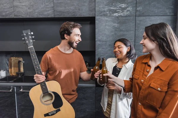 Homem alegre segurando guitarra acústica e clinking garrafas de cerveja com mulheres inter-raciais felizes na cozinha — Fotografia de Stock