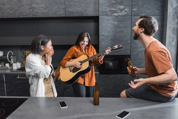 Возбужденная женщина с закрытыми глазами играет на акустической гитаре рядом с межрасовыми друзьями на кухне — стоковое фото