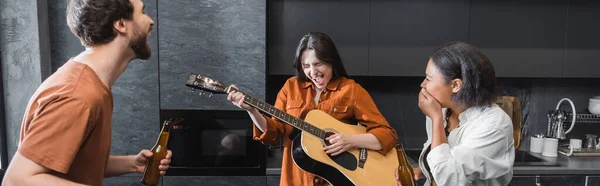 Excited woman with closed eyes playing acoustic guitar near interracial friends in kitchen, banner — Stock Photo