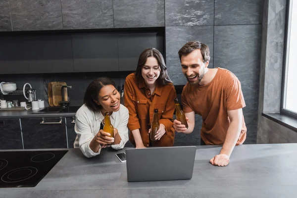 Amigos multiétnicos felices sosteniendo botellas de cerveza cerca de la computadora portátil mientras ve la película en la cocina - foto de stock