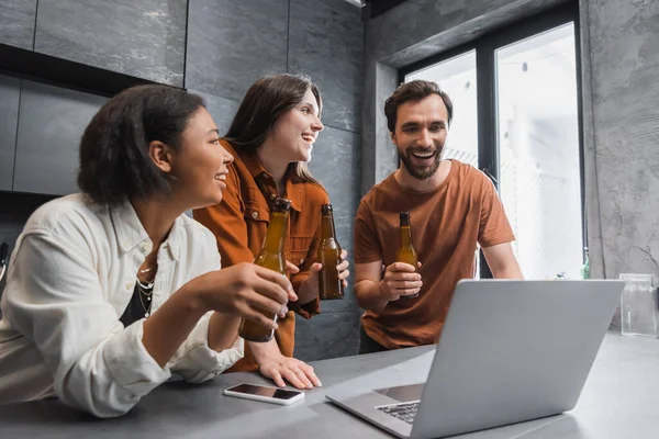 Happy multiethnic friends holding bottles of beer near laptop and smartphone with blank screen on kitchen worktop — Stock Photo