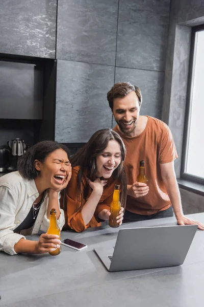Amigos multiétnicos felices sosteniendo botellas de cerveza y viendo la película de comedia en el portátil - foto de stock